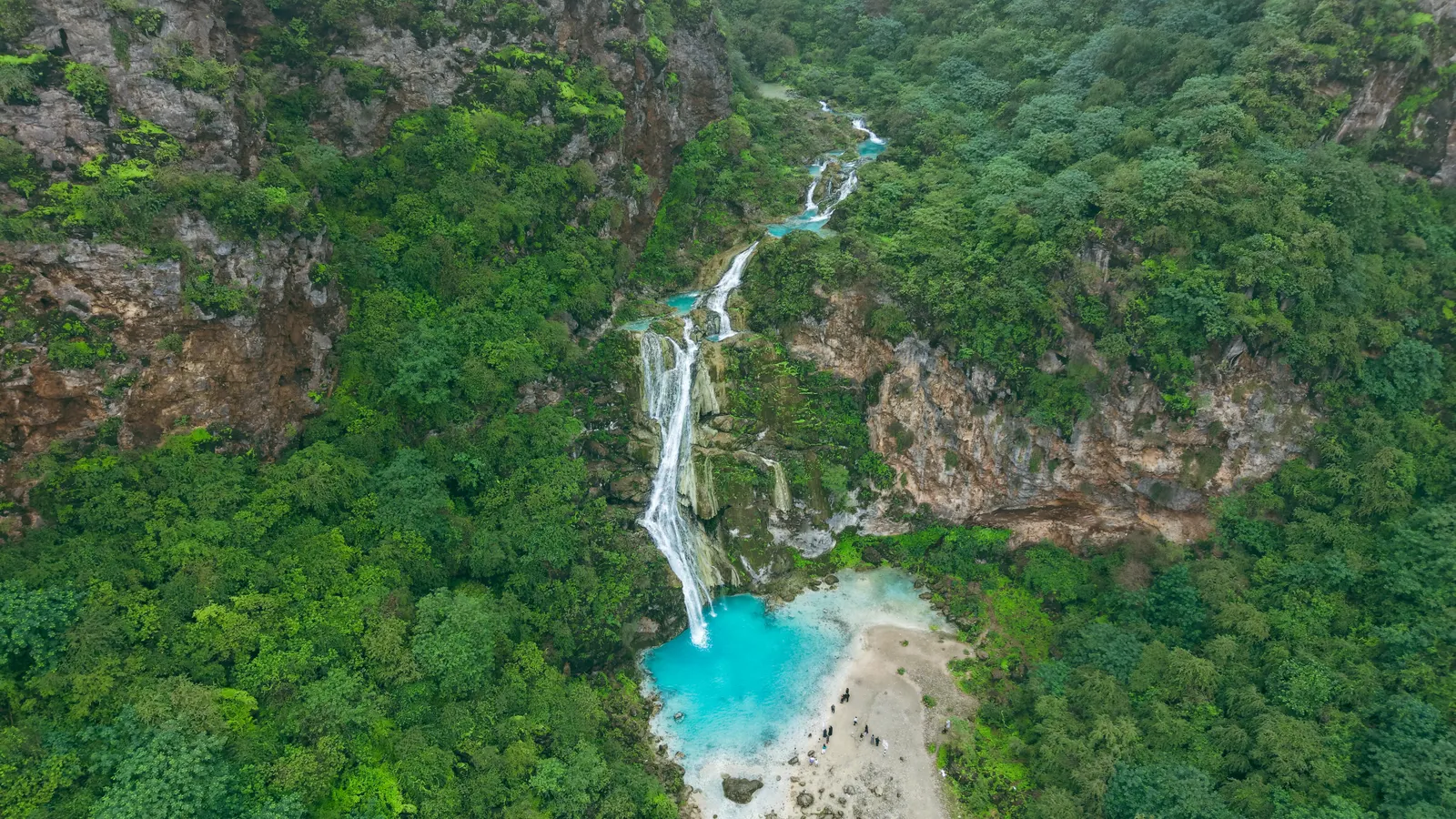 Aerial view of a seasonal waterfall falling into a turquoise pool between green, mist-covered mountains near Salalah during Khareef