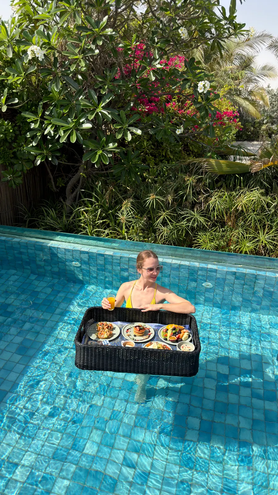 Floating breakfast tray of pancakes, pastries and fruit in the private pool of a villa at The Ritz-Carlton Ras Al Khaimah, Al Hamra Beach, with frangipani and bougainvillea behind