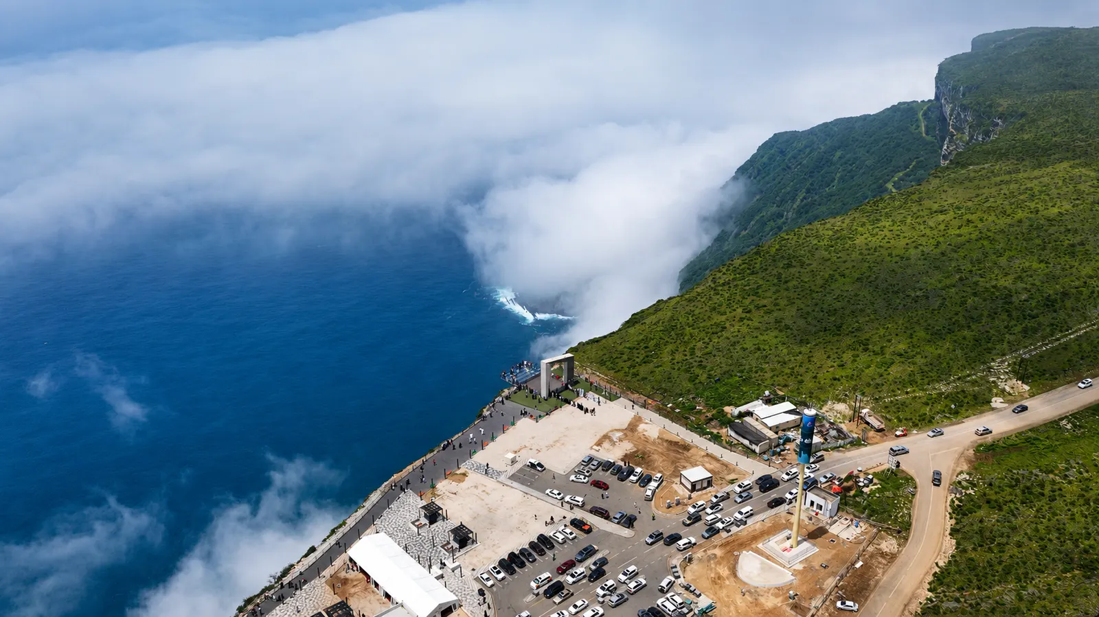 Cloud pouring over a green clifftop above the sea at a coastal viewpoint near Salalah, with a car park and visitors on the headland below